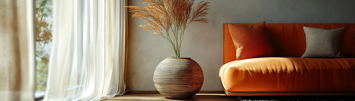 Orange sofa with decorative pillows next to a vase with dried plants on a windowsill.